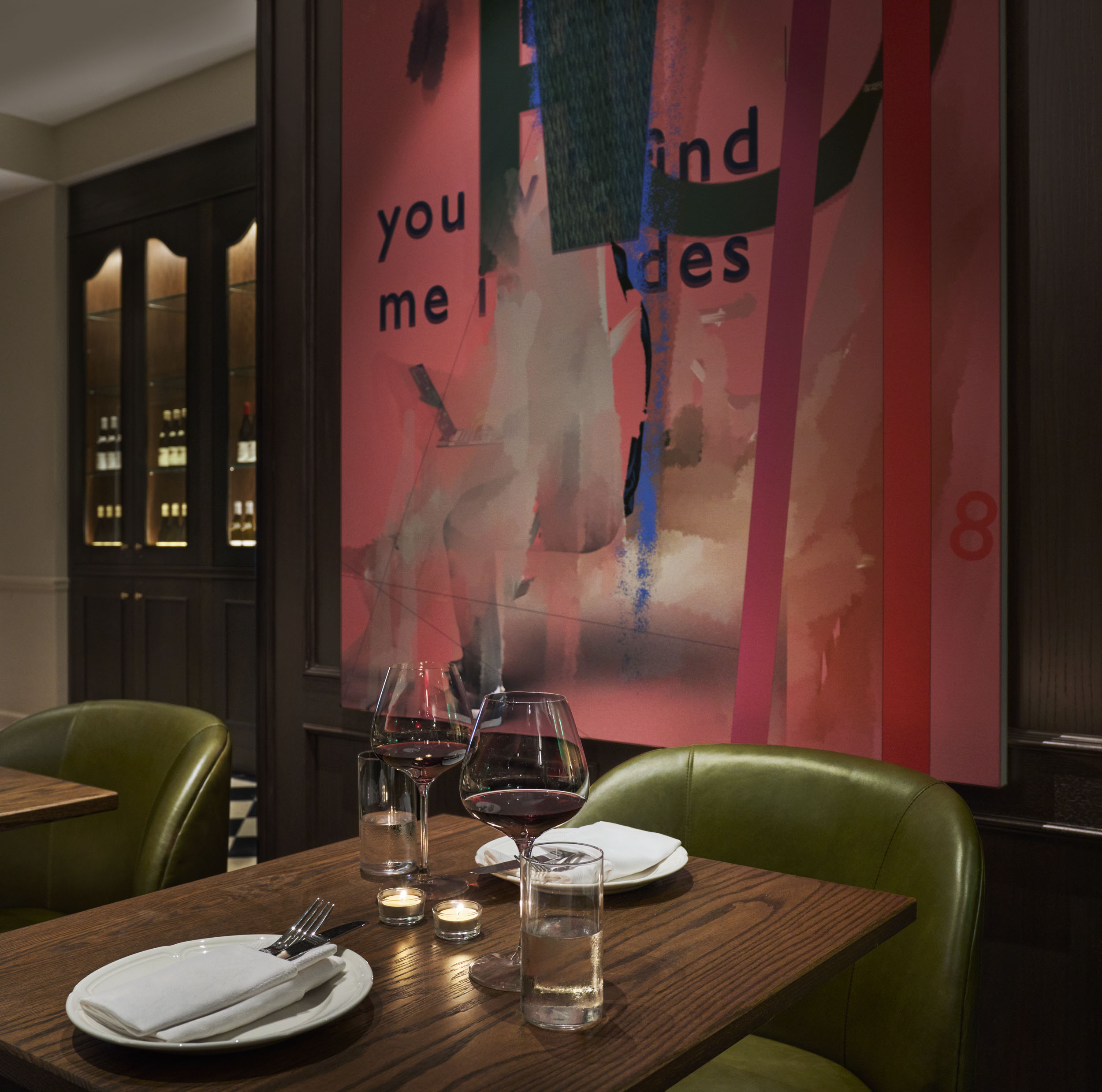 A dining area with a wooden table set for two, green leather chairs, wine glasses, and candle lights, in front of a large abstract artwork.