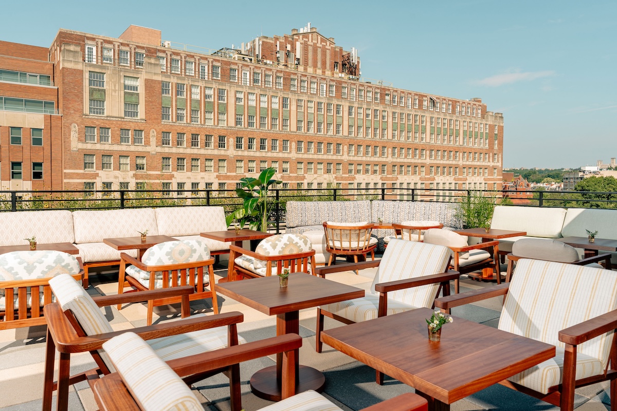 A rooftop outdoor seating area with wooden tables and chairs, and white cushions, overlooking a large brick building.