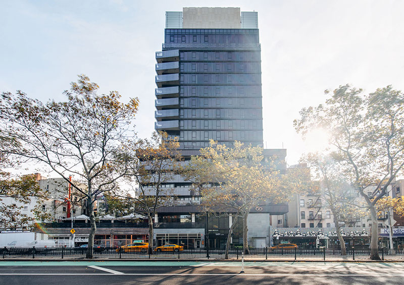 A high-rise building with a glass facade stands tall on a city street corner, surrounded by trees and urban landscape.