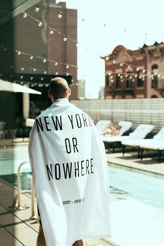 A man stands by a pool with a city view, wearing a robe that says 'New York or Nowhere'.