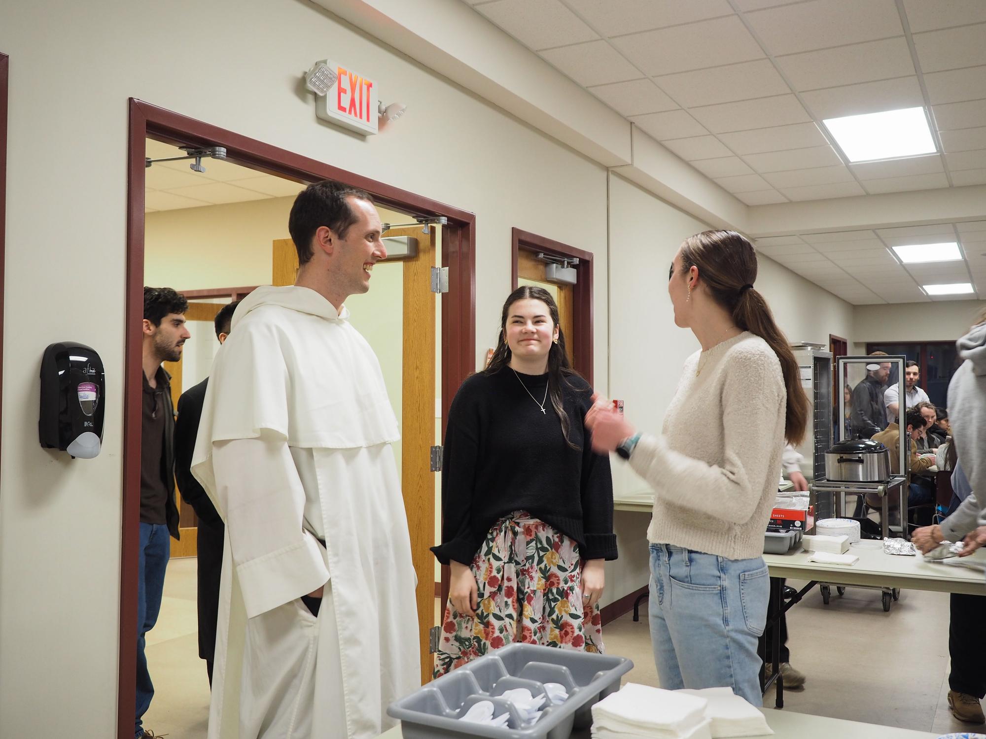 A priest in white vestments
							  smiles and talks to young adults in a large room.
