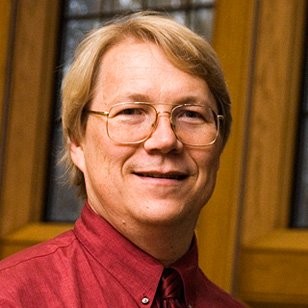 A man in a red collared shirt and wire-rimmed glasses smiles in a wood-paneled room 