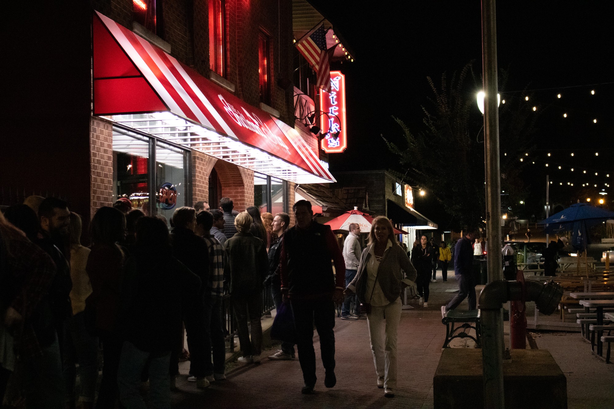 A line of people outside Nick's and people walking on the sidewalk adjacent to Kirkwood Avenue.