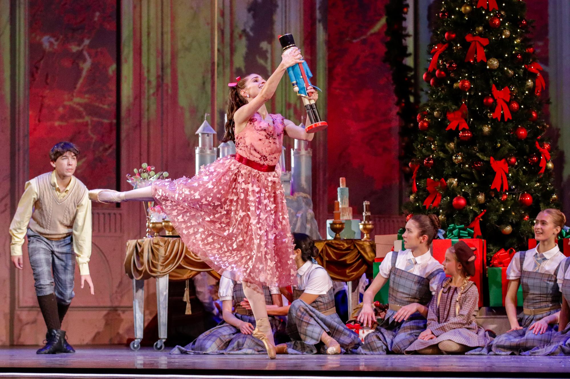 A dancer in a pink dress dances in front of a Christmas tree with a nutcracker doll as children onstage look on