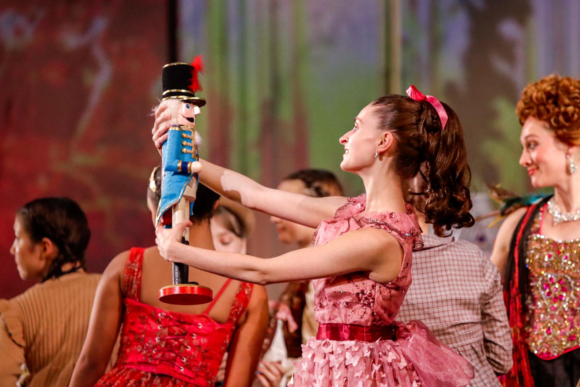 A dancer in a pink dress holds the blue nutcracker doll aloft.