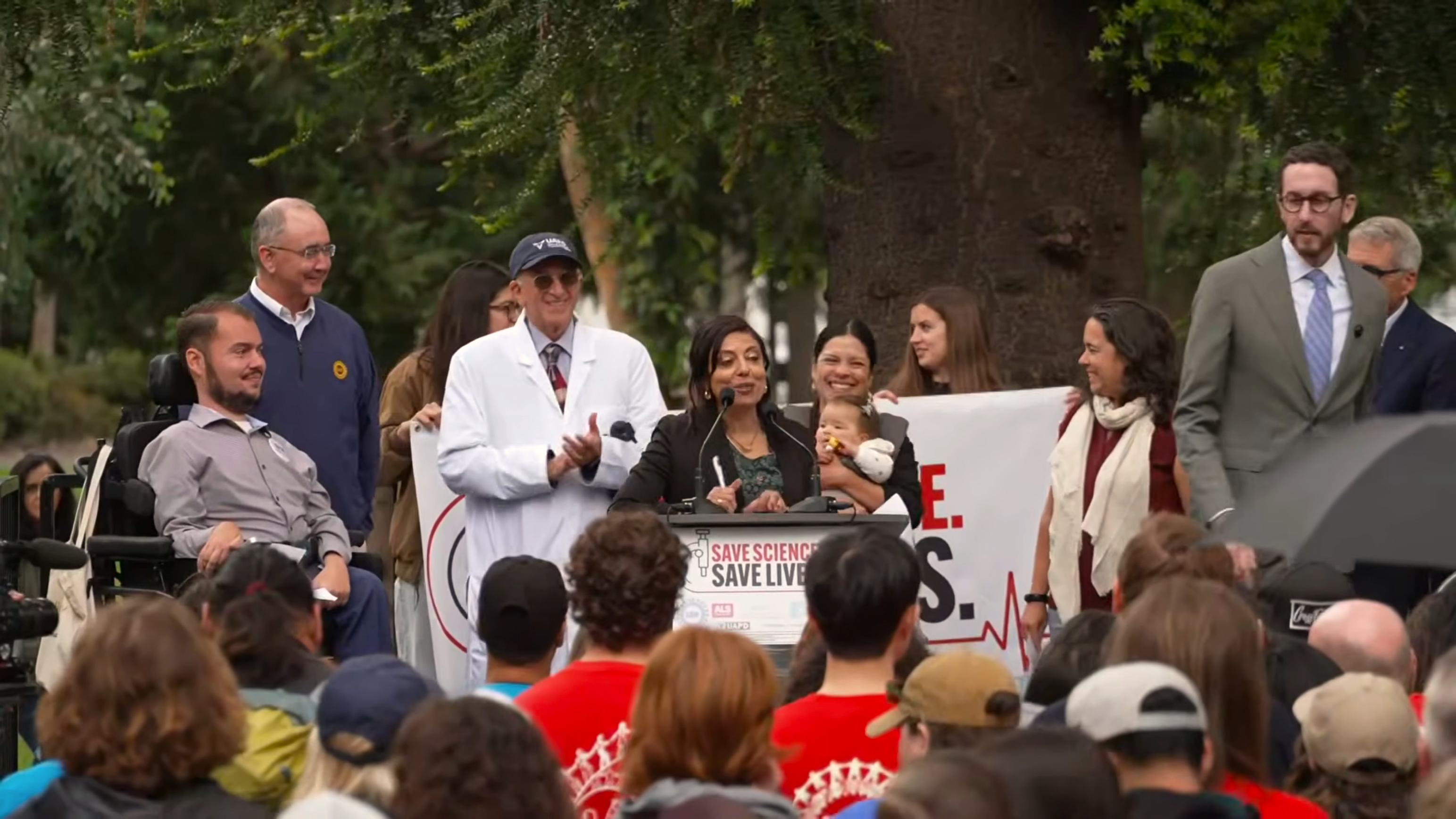 Professor Monica Gandhi speaking on a podium in front of a large crowd with plackards and signs.