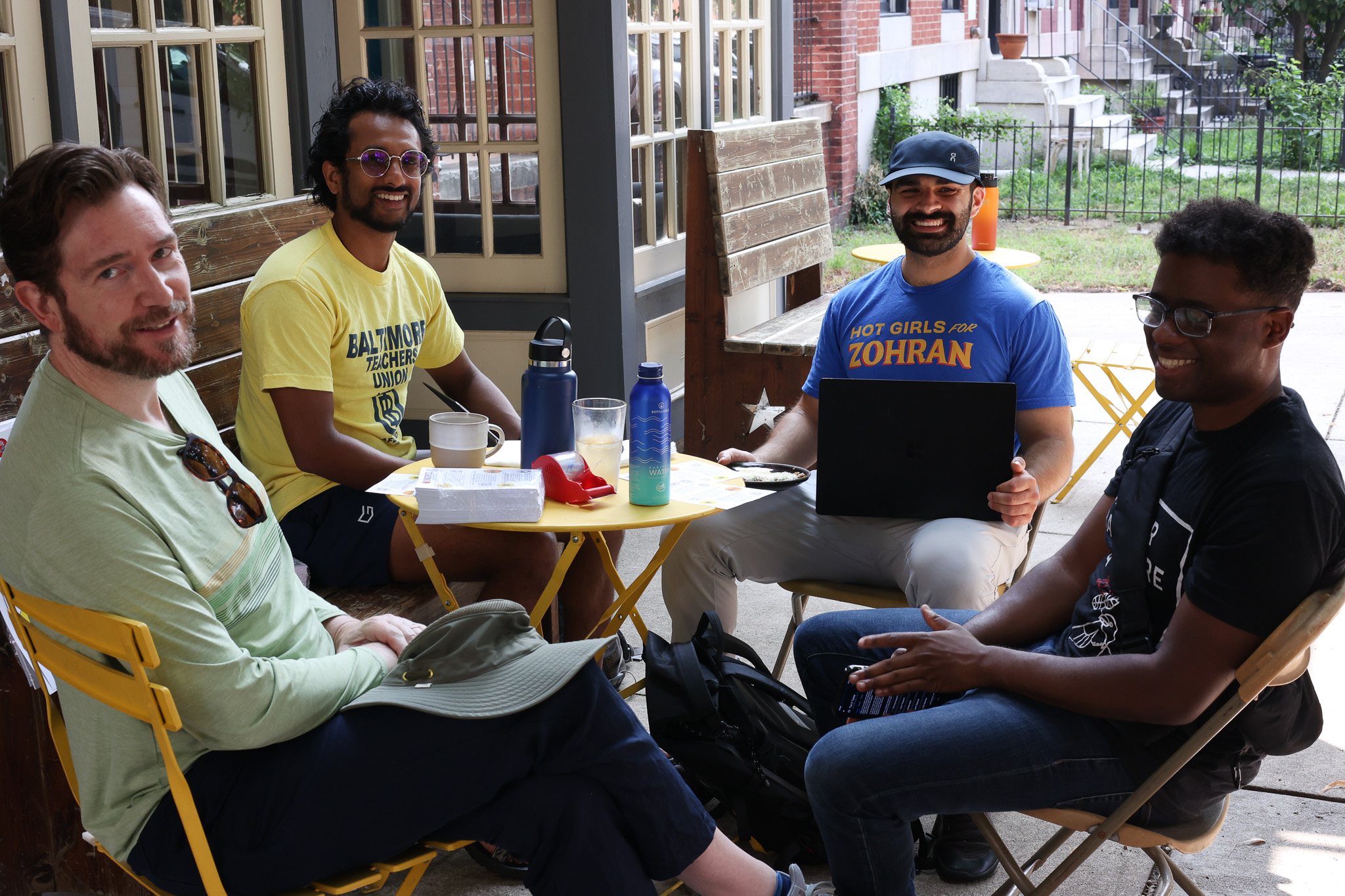 Four GBDSA members at a small cafe table outside. One is wearing a Baltimore Teachers Union shirt, and another is wearing a Hot Girls for Zohran shirt.