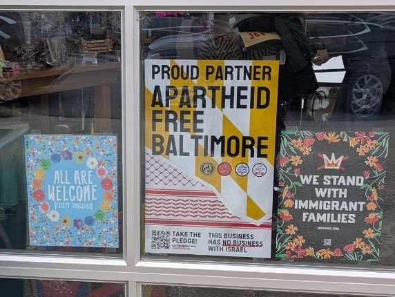 Poster in store-front for Apartheid-Free Baltimore. Proud Partner Apartheid Free Baltimore. This business has no business with Israel. Take the pledge! Accompanying posters in storefront for "We stand with immigrant families" and "all are welcome (except fascists)".