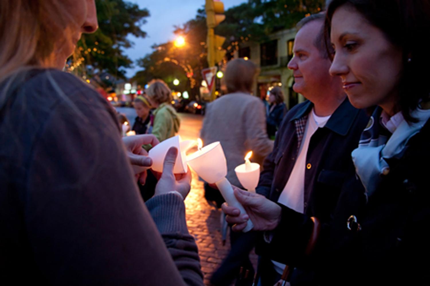 People gather at dusk for a candlelight vigil, holding lit candles along a city street.