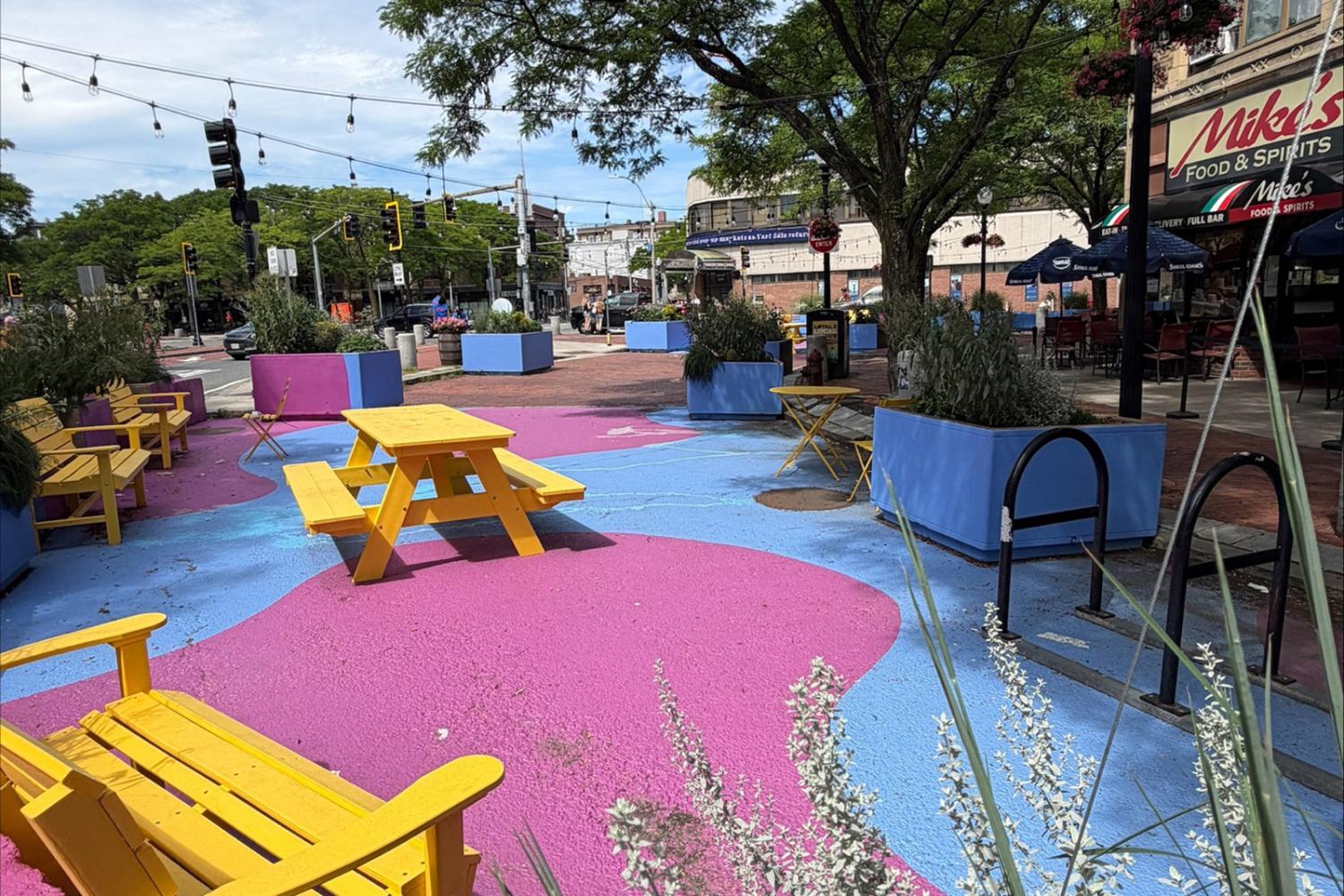 Davis Square plaza with pink and blue pavement, yellow seating, blue planters, string lights, and mature trees.