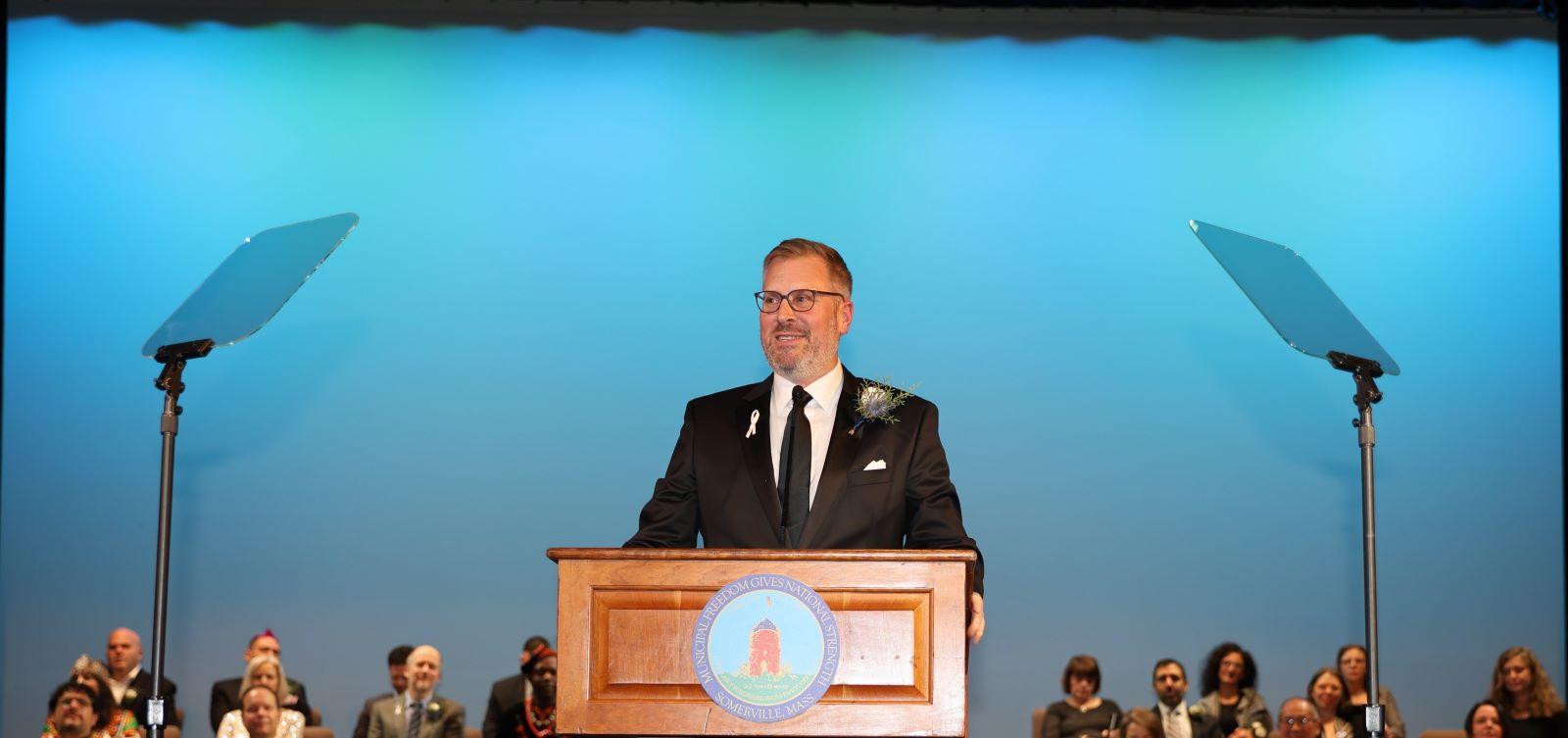 Mayor Jake Wilson delivers his inaugural address on stage in front of a blue background. 