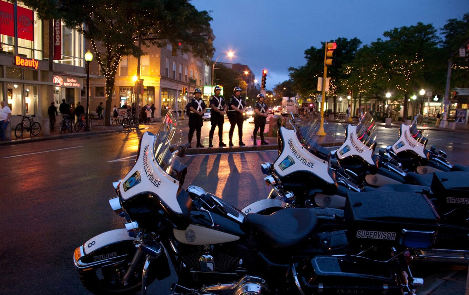 Four Somerville Police officers stand solemnly in front of a row of SPD motorcycles during a 9/11 vigil in Davis Sq. at night time