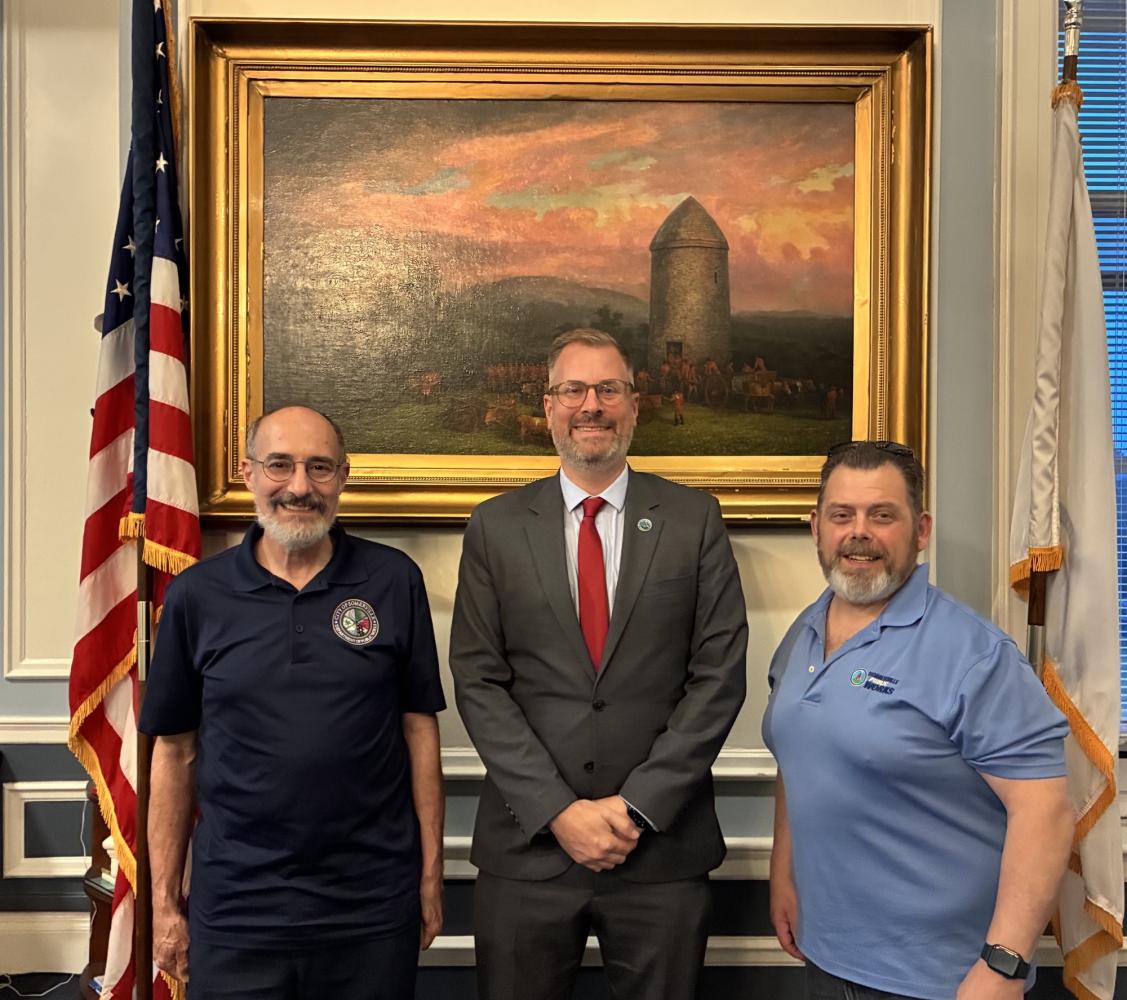 Three men, including Mayor Jake Wilson, in front of a painting with an American flag. 