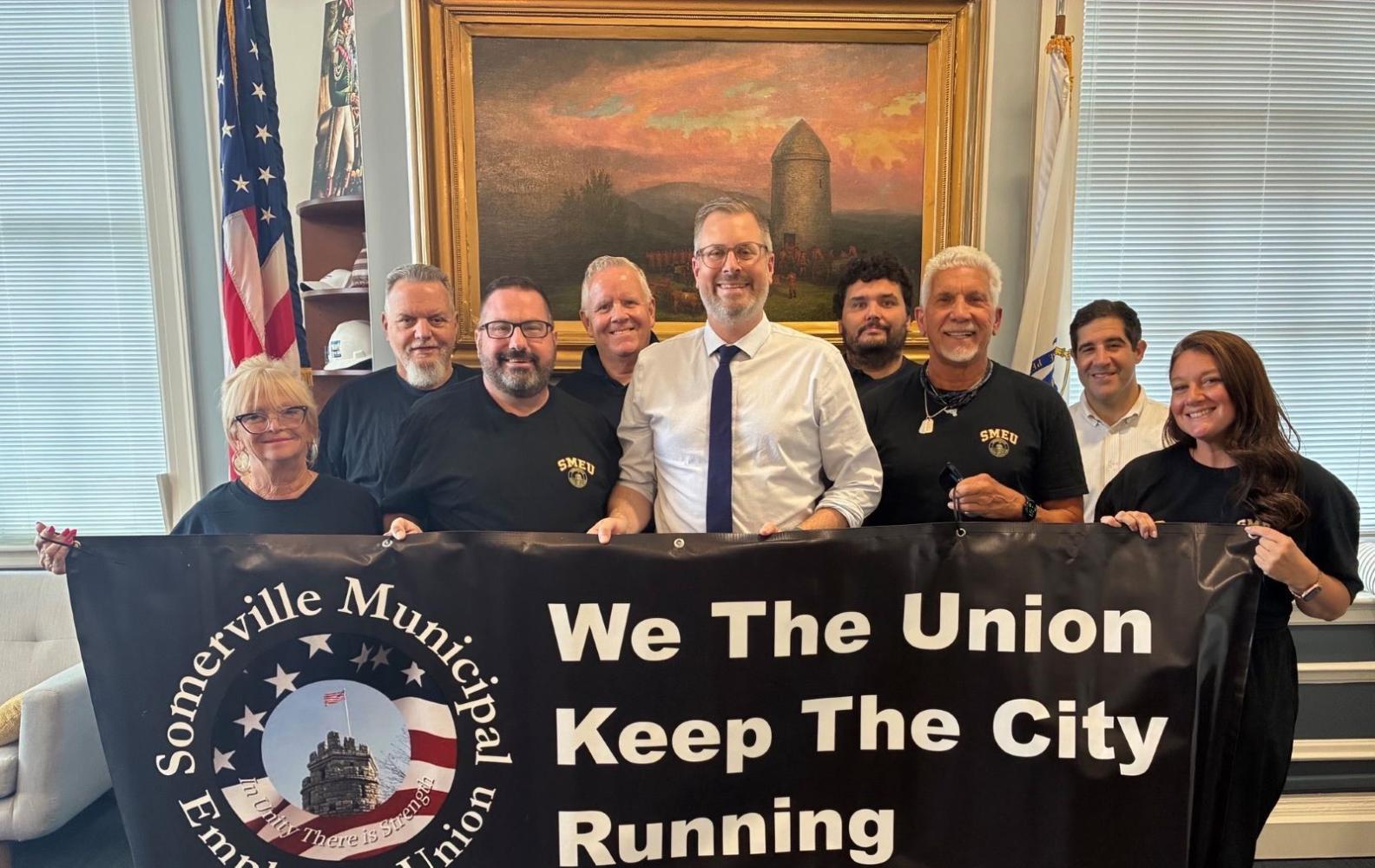 Members of the SMEU Unit A stand with Mayor Jake Wilson in front of a banner saying "We the union keep the city running.'