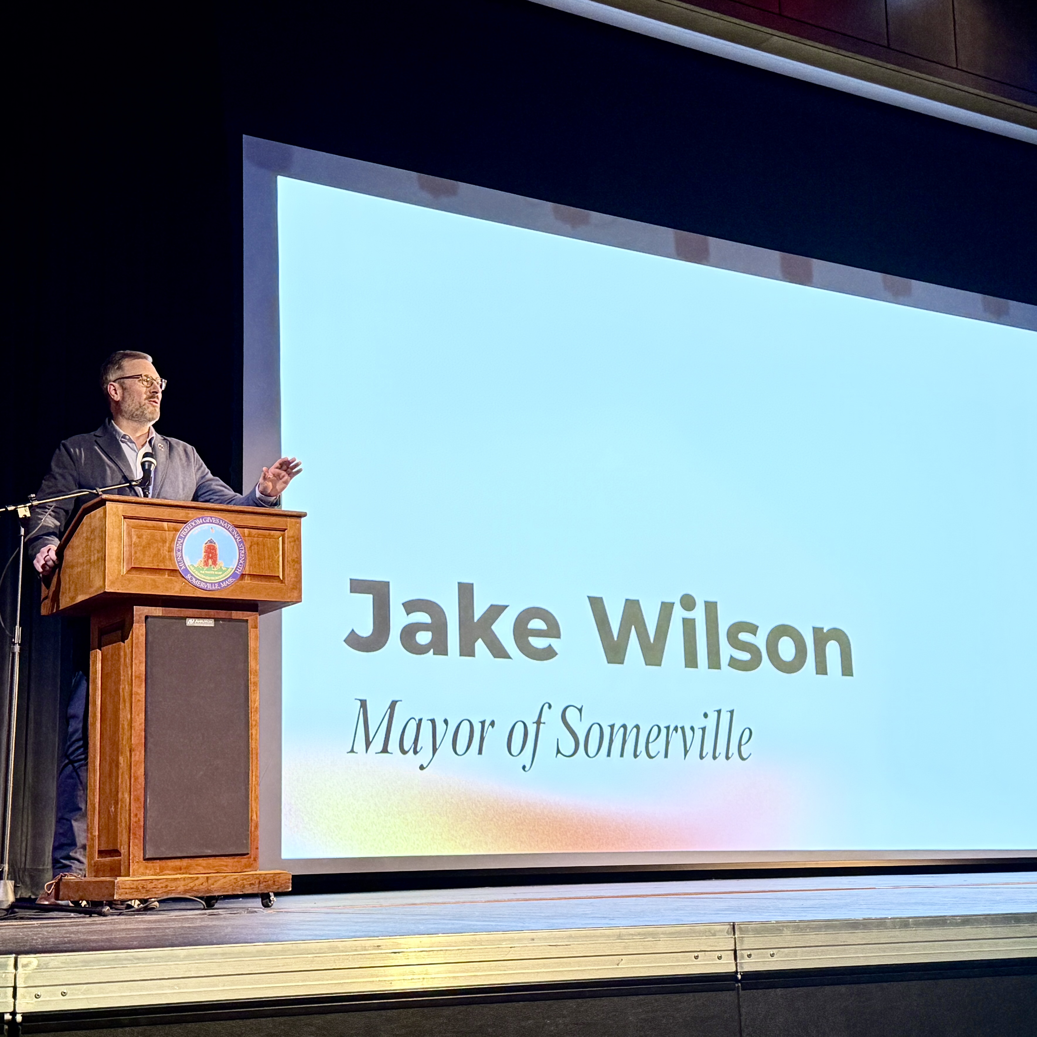 Mayor Jake Wilson at a podium presenting in front of a screen on stage. 