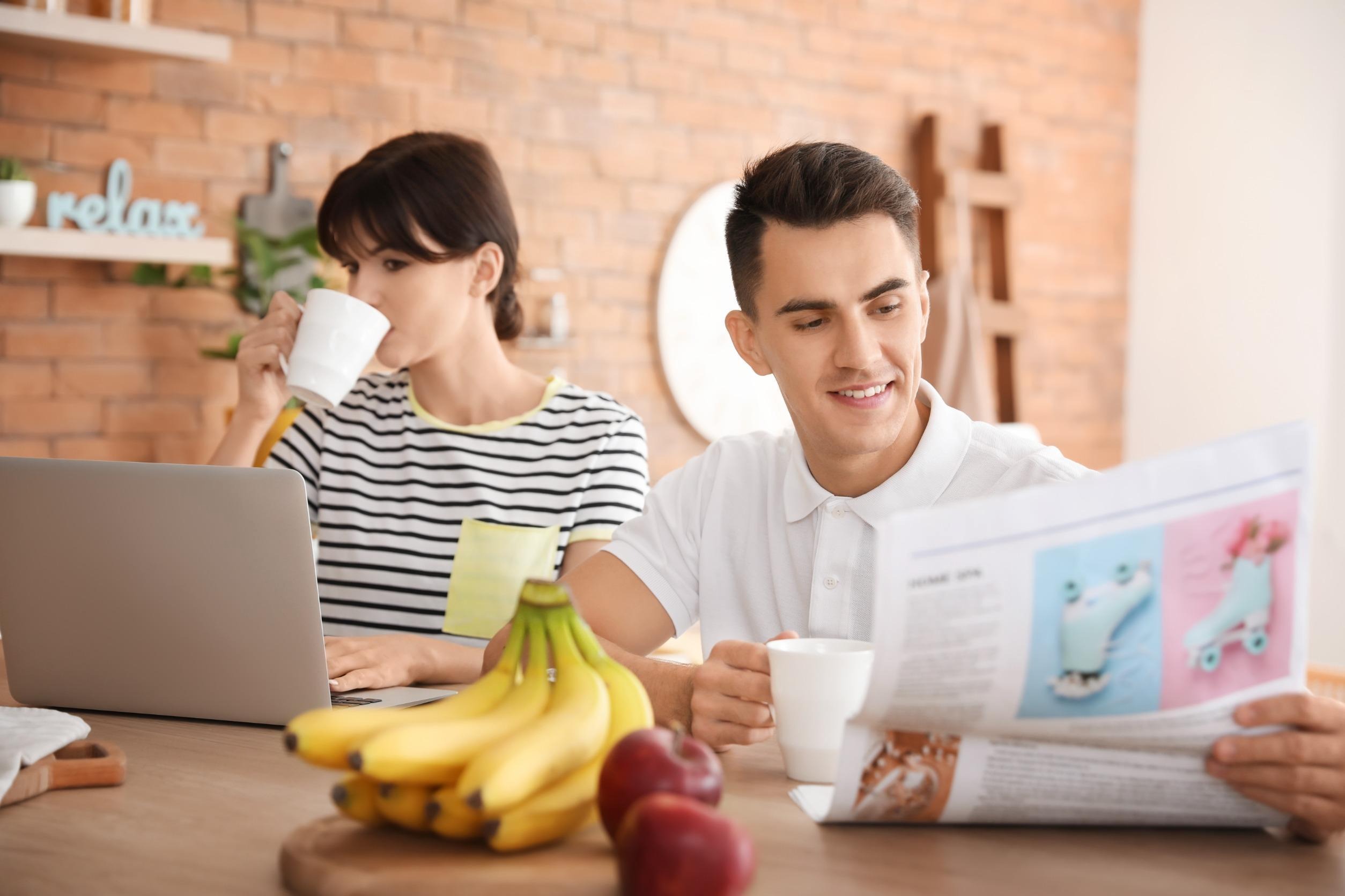 woman looking at laptop, man reading newspaper, both drinking coffee