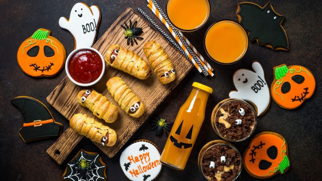 Flat lay of various Halloween treats including cookies, juice, and mummy hot dogs