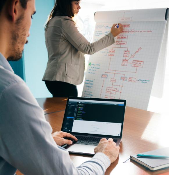 a business woman draws on a white board while a man checks his laptop screen at a desk in front of her, you are looking over his shoulder at what she is writing.