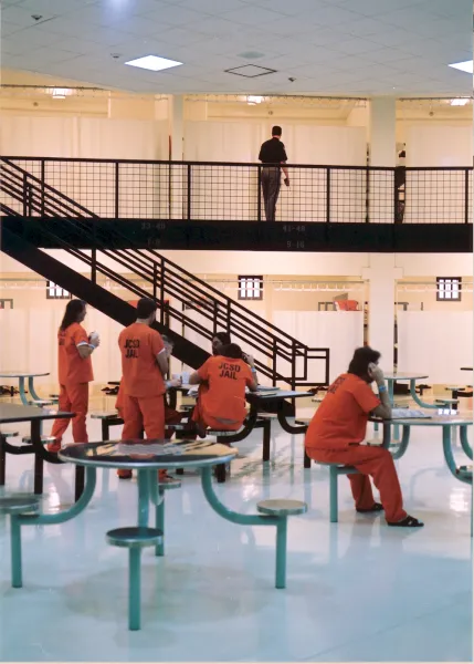 several inmates sit at tables in their pod while a corrections officer walks the second floor