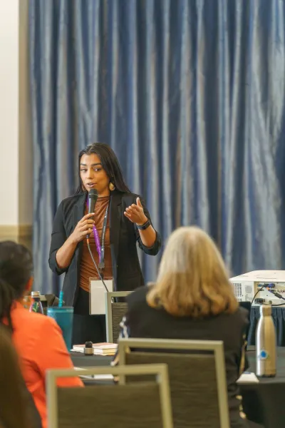 A woman speaks in front of a crowd of other women at a conference with a blue curtain behind her