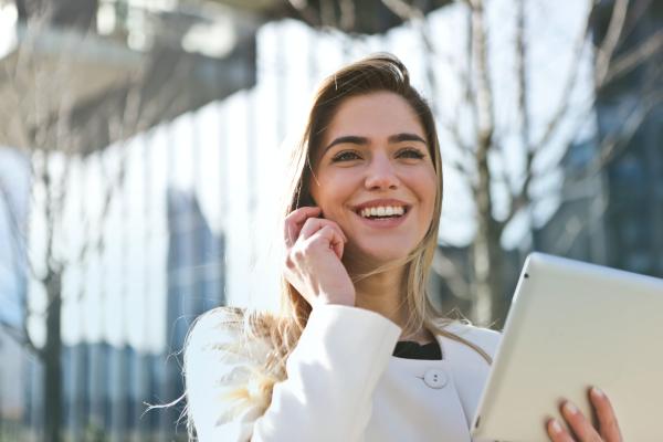 A smiling woman talking on a cell phone while holding a tablet in a city setting