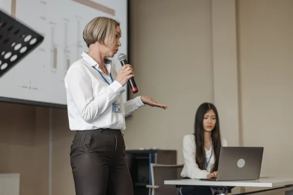 a woman speaking in front of a projected slide, meanwhile another woman works on a laptop off to the side