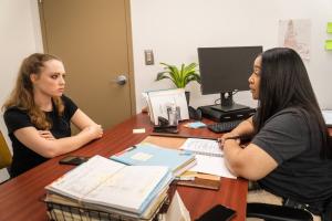 Probation office, a probation officer interviews a woman across an office desk filled with books and papers
