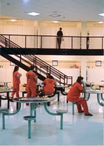 several inmates sit at tables in their pod while a corrections officer walks the second floor