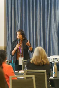 A woman speaks in front of a crowd of other women at a conference with a blue curtain behind her