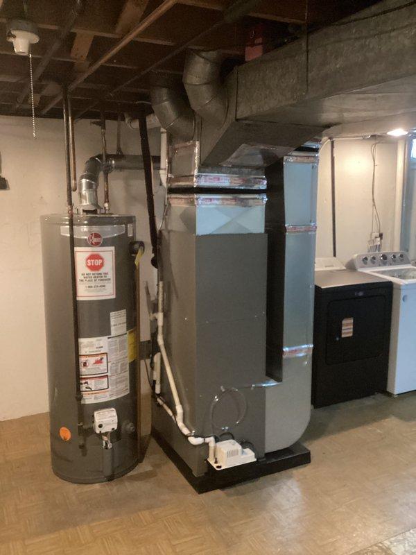 # HVAC Complete System Installation

Successfully completed installation of a new 70,000 BTU furnace and 2-ton air conditioning system in basement utility area with left-side configuration. Installed gray metal air handler with proper ductwork connections, including metal ductwork extending vertically with secure tape sealing at all joints. Mounted white condensate pump on black drain pan at base of air handler with clear tubing for proper drainage. Reconnected all necessary plumbing, electrical, and control wiring to integrate new system with existing infrastructure. All equipment positioned to allow adequate clearance and service access alongside existing water heater. System tested and operational upon completion.