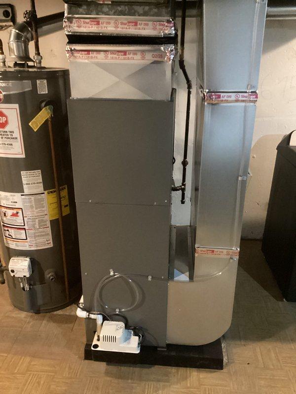 # HVAC Complete System Installation

Successfully completed installation of a new 70,000 BTU furnace and 2-ton air conditioning system in basement utility area with left-side configuration. Installed gray metal air handler with proper ductwork connections, including metal ductwork extending vertically with secure tape sealing at all joints. Mounted white condensate pump on black drain pan at base of air handler with clear tubing for proper drainage. Reconnected all necessary plumbing, electrical, and control wiring to integrate new system with existing infrastructure. All equipment positioned to allow adequate clearance and service access alongside existing water heater. System tested and operational upon completion.