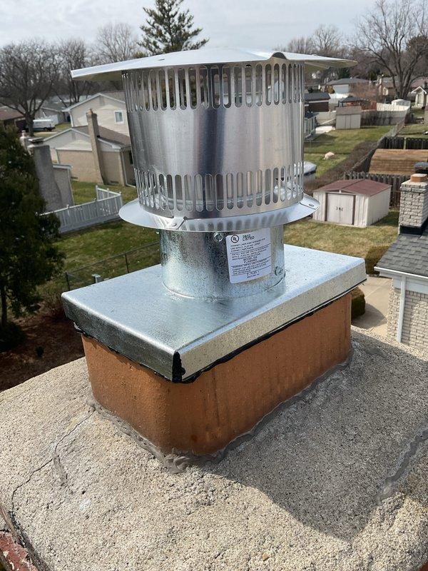 Completed return visit to install rain cap assembly on existing chimney liner system. Mounted galvanized metal rain cap with conical design onto cylindrical chase cover positioned on brick chimney structure, securing all components to prevent water intrusion while maintaining proper ventilation for exhaust gases.