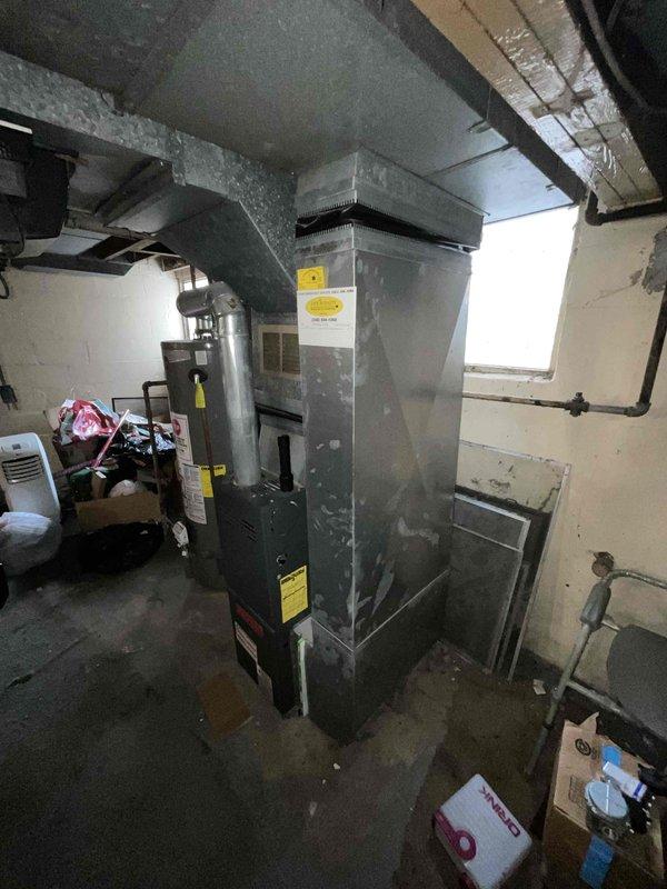 Conducted furnace replacement estimate for non-operational natural gas heating system located in basement with existing galvanized ductwork and exposed joists. Assessed current gray metal furnace unit with multiple warning labels, evaluated existing supply and return duct configuration including vertical galvanized trunk line with elbow connection, and documented utility room conditions including concrete block walls, copper gas piping with brass fittings, and available clearances for new equipment installation.
