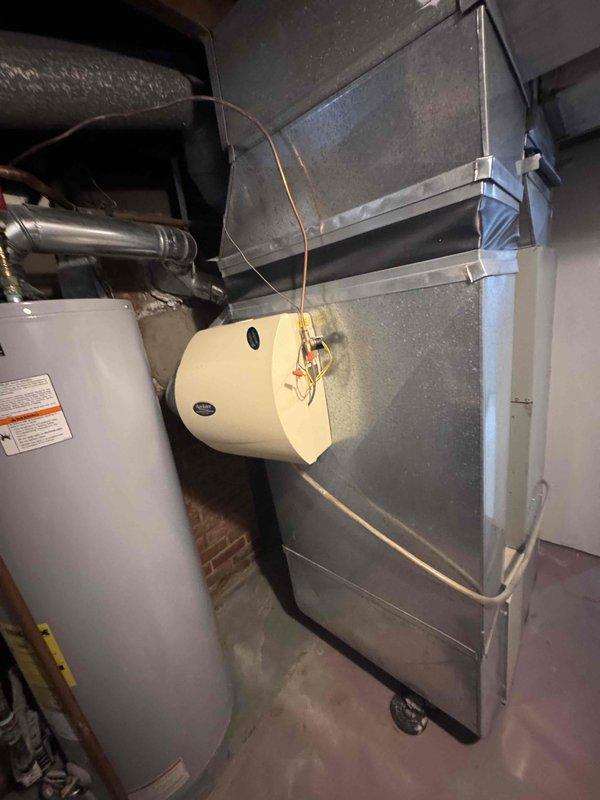 Completed comprehensive HVAC system evaluation for 20-year-old natural gas furnace and air conditioning equipment located in basement. Assessed existing blower motor assembly showing significant age-related wear, moisture damage, and corrosion on metal components and surrounding concrete base with visible efflorescence and deterioration. Documented current ductwork configuration including humidifier accessory unit mounted to main supply plenum, water heater proximity, and overall mechanical room layout to provide accurate replacement system specifications and installation requirements.