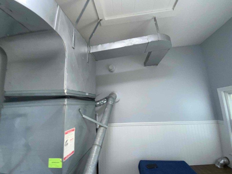 Conducted on-site evaluation for central air conditioning installation in residential property with no existing central air system. Assessed existing HVAC infrastructure including furnace/air handler unit, ductwork configuration, and mechanical space in basement utility room. Evaluated gray metal ductwork system, flexible ducting connections, and vertical cylindrical duct routing for compatibility with proposed AC system integration. Documented current equipment setup, including air handler positioning, duct runs along ceiling, and available space for outdoor condenser unit placement. Photographed existing installation to support estimate preparation for complete central air conditioning system addition scheduled for spring installation.