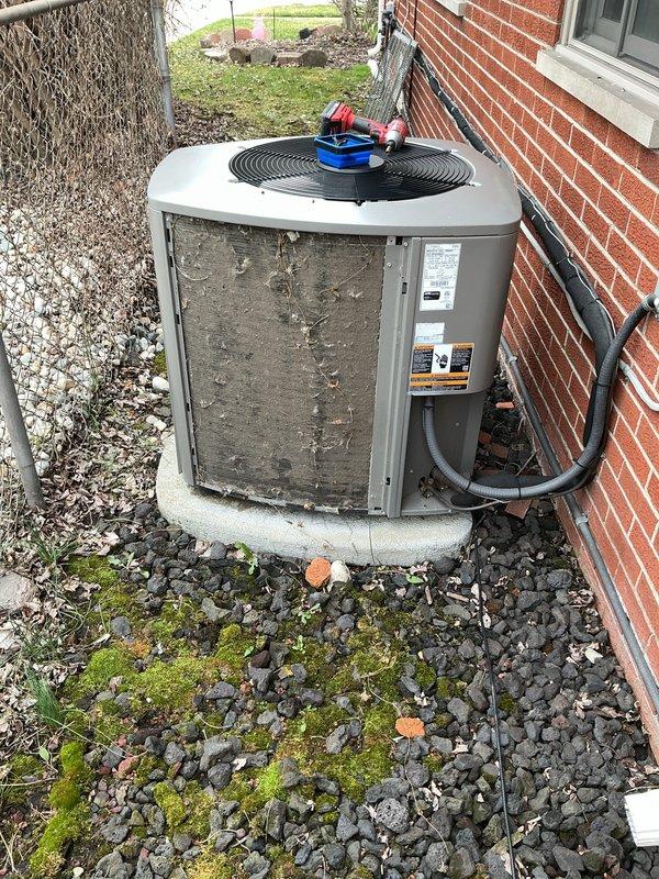 Performed comprehensive air conditioning maintenance on residential outdoor condenser unit. Conducted visual inspection of unit exterior, verifying proper placement on concrete pad and checking for physical damage or deterioration. Inspected electrical conduit connections and verified secure attachment to building structure. 

Removed debris, leaves, and organic material from surrounding gravel base and unit perimeter to ensure adequate airflow and drainage. Noted moss growth in gravel bed indicating moisture retention in area. Checked manufacturer labels and unit specifications for proper identification and maintenance requirements.

Assessed top-mounted fan assembly and protective grille for structural integrity and proper operation. Utilized diagnostic gauges to measure system pressures and verify refrigerant charge levels. Inspected condenser coil fins through louver openings for blockage, damage, or excessive dirt accumulation. Verified unit levelness on pad and checked for signs 