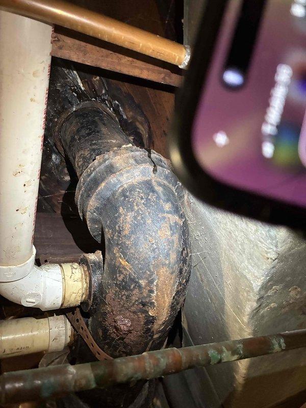 Responded to a plumbing emergency involving a leaking cast iron pipe connected to the toilet drainage system. Upon inspection, discovered severe corrosion and surface deterioration on the cast iron waste pipe, with heavy oxidation, rust patches, and flaking material indicating advanced degradation of the pipe structure. The corroded section showed active deterioration with significant structural compromise, particularly around curved fittings where moisture exposure had accelerated the breakdown of the cast iron material. Documented the extensive damage with photographs showing the contrast between the failing cast iron components and adjacent PVC and copper piping in serviceable condition. Assessed the confined access area beneath the structure and identified the scope of repair work required to address the leak source and prevent further water damage.