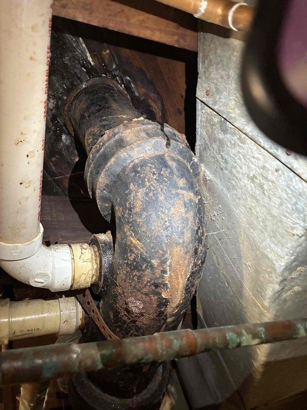 Responded to a plumbing emergency involving a leaking cast iron pipe connected to the toilet drainage system. Upon inspection, discovered severe corrosion and surface deterioration on the cast iron waste pipe, with heavy oxidation, rust patches, and flaking material indicating advanced degradation of the pipe structure. The corroded section showed active deterioration with significant structural compromise, particularly around curved fittings where moisture exposure had accelerated the breakdown of the cast iron material. Documented the extensive damage with photographs showing the contrast between the failing cast iron components and adjacent PVC and copper piping in serviceable condition. Assessed the confined access area beneath the structure and identified the scope of repair work required to address the leak source and prevent further water damage.