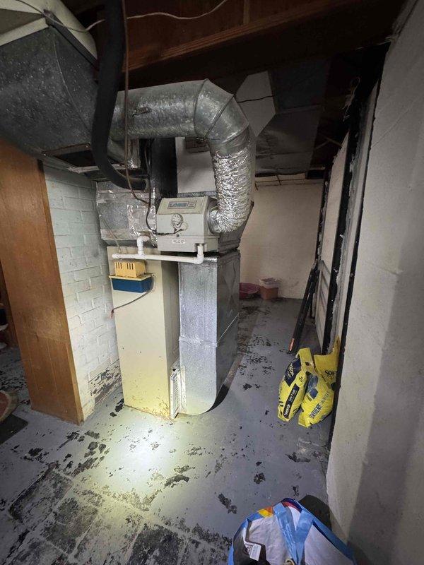 Conducted on-site evaluation for furnace replacement at residential property with 40-year-old non-operational heating system. Inspected existing cream-colored furnace unit with gray upper section located in basement utility area, noting complete system failure and age-related deterioration. Assessed existing ductwork configuration including flexible metallic ducts and rigid metal components connected to ceiling joists. Evaluated electrical infrastructure including main panel box and wiring capacity for new equipment installation. Documented basement mechanical room conditions, concrete block wall structure, floor joists, and available clearances for replacement unit. Provided comprehensive replacement estimate for new furnace system to restore heating functionality.