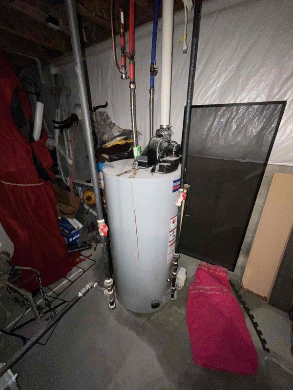 Responded to water heater leak in basement utility area. Inspected white cylindrical tank water heater with multiple supply line connections, confirming active leak condition. Documented existing plumbing configuration including hot and cold water lines, pressure relief valve assembly, and top-mounted circulation pump. Photographed unit condition, piping connections, and surrounding mechanical systems to provide comprehensive estimate for water heater replacement. Customer previously had HVAC system installed in 2015.