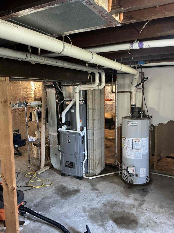 Conducted on-site assessment for gas water heater replacement in 1000 square foot residential property with walk-in basement access. Evaluated existing white cylindrical gas-fired water heater unit positioned adjacent to HVAC system, documenting current copper supply line connections, gas hookup configuration, and venting requirements. Photographed installation area including exposed ceiling joists, concrete floor conditions, existing plumbing infrastructure with overhead PVC drainage system, and clearance space available for new unit installation to provide accurate replacement quote.