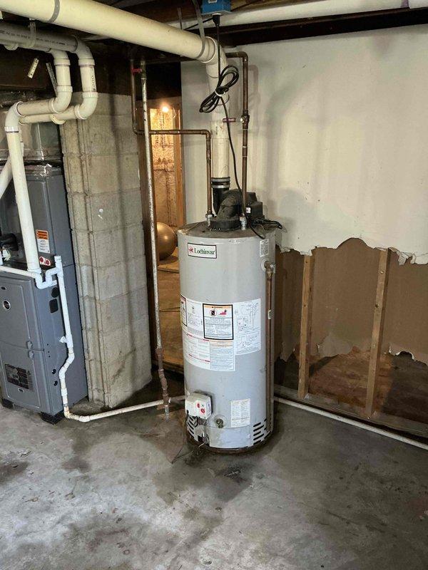 Conducted on-site assessment for gas water heater replacement in 1000 square foot residential property with walk-in basement access. Evaluated existing white cylindrical gas-fired water heater unit positioned adjacent to HVAC system, documenting current copper supply line connections, gas hookup configuration, and venting requirements. Photographed installation area including exposed ceiling joists, concrete floor conditions, existing plumbing infrastructure with overhead PVC drainage system, and clearance space available for new unit installation to provide accurate replacement quote.