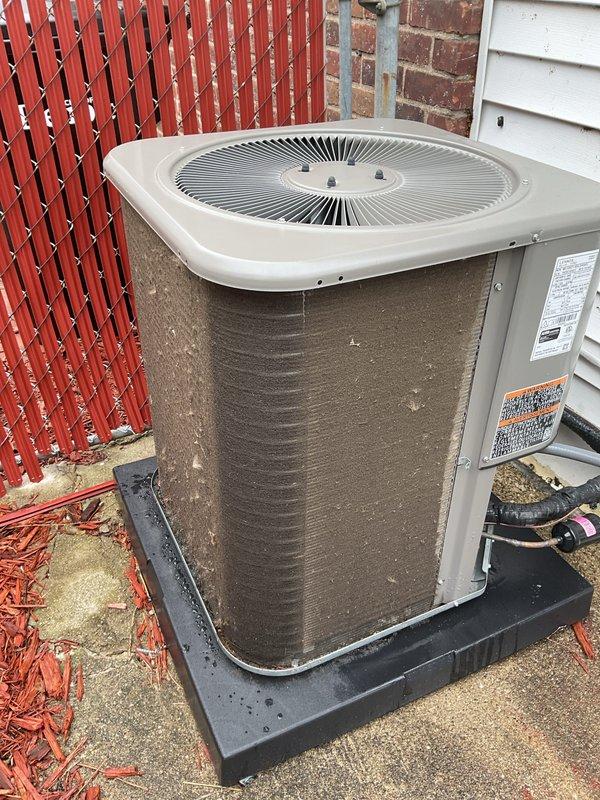 Completed scheduled air conditioning maintenance on residential outdoor condenser unit. Performed comprehensive inspection of unit positioned on concrete pad against building exterior, verified proper operation of fan assembly and circular grille protection system, checked manufacturer labels and safety markings for compliance, and confirmed refrigerant lines and electrical conduit connections were secure. Unit found in good functional condition with no visible damage, excessive wear, or deterioration requiring immediate attention.