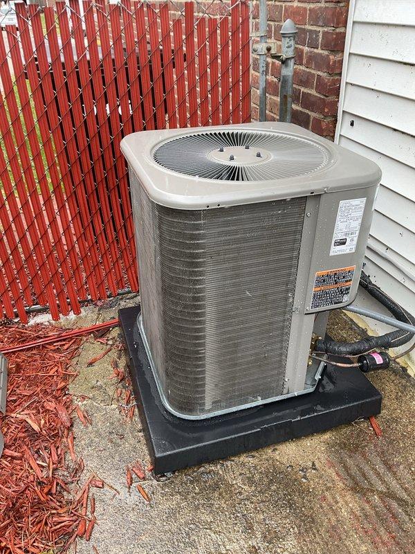 Completed scheduled air conditioning maintenance on residential outdoor condenser unit. Performed comprehensive inspection of unit positioned on concrete pad against building exterior, verified proper operation of fan assembly and circular grille protection system, checked manufacturer labels and safety markings for compliance, and confirmed refrigerant lines and electrical conduit connections were secure. Unit found in good functional condition with no visible damage, excessive wear, or deterioration requiring immediate attention.