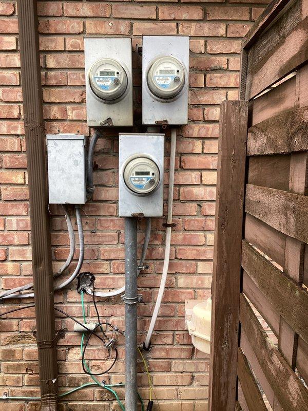 Completed estimate for whole-home surge protector installation at a 1972 condominium. Inspected existing electrical service consisting of multiple digital electric meters mounted on exterior brick wall with weathered metal enclosures. Documented current metering configuration showing three OpenWay digital meters serving multiple units with various conduits and wiring connections. Assessed panel accessibility and existing grounding system, noting exposed grounding conductors present at installation. Photographed meter bank arrangement and service entrance equipment to determine optimal surge protection device placement and compatibility with existing infrastructure. Provided recommendations for Type 2 surge protective device installation based on observed electrical service configuration and building age.