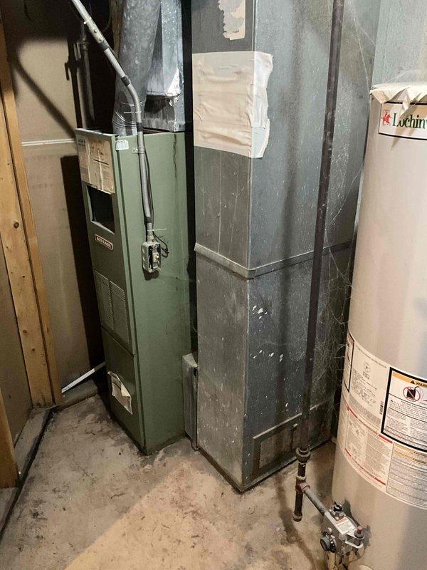 Conducted on-site evaluation for complete HVAC system replacement in residential side-by-side condo unit. Assessed existing olive-green furnace located in basement mechanical room with connected ductwork assembly showing age-related wear and surface discoloration. Evaluated outdoor condensing unit positioned on concrete pad against exterior wall, noting weathered white/beige cabinet with circular top-discharge fan grille, flexible conduit electrical connection, and proximity to PVC plumbing configuration. Documented current equipment conditions through photographic inspection to provide comprehensive replacement estimate for furnace and air conditioning system planned for installation within upcoming two-week timeframe.