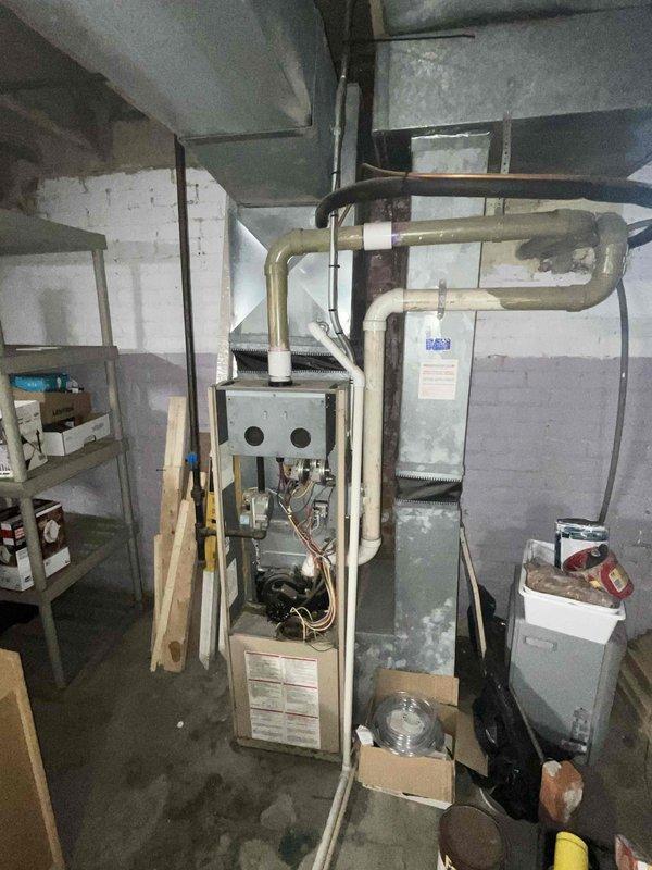 Completed comprehensive HVAC system evaluation for full system replacement estimate in basement utility area. Documented existing aging furnace unit showing significant wear, paint deterioration, and dust accumulation, along with connected ductwork and associated mechanical components. Photographed current system configuration including furnace with exposed burner assembly, blower motor, wiring connections, and galvanized metal ductwork to provide detailed assessment for complete system replacement proposal.
