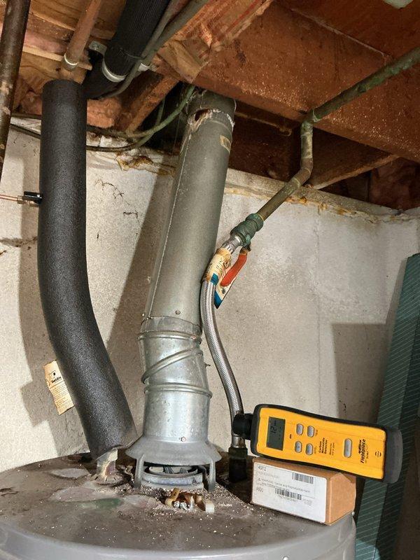 Completed annual HVAC maintenance inspection on water heater system. Performed diagnostic testing using digital meter to verify proper combustion and system operation, inspected flue pipe and venting components for proper draft and clearances, and examined all connections including gas flex line and associated fittings. Documented current condition of unit showing normal wear and debris accumulation at base, confirmed all safety controls functioning within specifications.