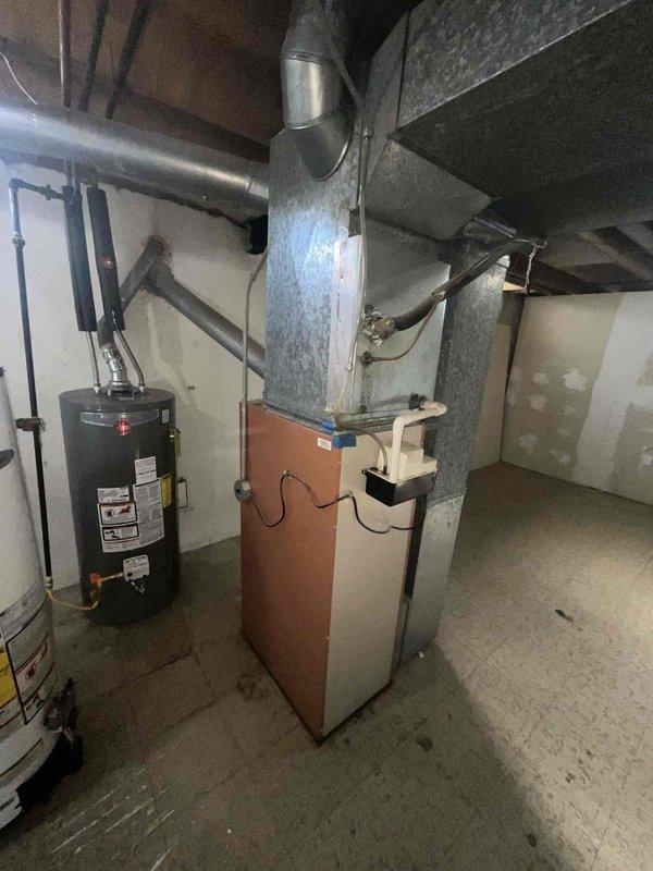 Conducted comprehensive site evaluation for complete HVAC system replacement at residential property. Existing 16-year-old air conditioning unit experiencing failure, blowing hot air instead of cooling. Assessed basement mechanical room containing aging furnace with two-tone exterior casing and cylindrical water heater. Documented existing ductwork configuration including large galvanized metal supply and return ducts running horizontally across exposed ceiling joists. Evaluated current system layout, clearances, and installation access points for full system replacement. Photographed equipment positioning, duct connections, electrical service, and mechanical room infrastructure to prepare detailed replacement proposal. Provided comprehensive estimate for new HVAC system installation to address immediate cooling failure and aging equipment concerns.