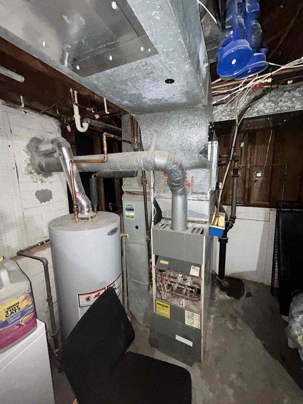 Conducted comprehensive HVAC system evaluation for furnace and air conditioning replacement estimate at lower tri-level residence with 18-year-old existing natural gas heating and cooling equipment currently in operational condition. Assessed existing furnace unit located in basement mechanical room, documenting current system configuration including ductwork layout, venting arrangements, and electrical connections. Evaluated air conditioning system components and refrigerant line routing. Photographed existing HVAC installation showing furnace cabinet with control panels, extensive galvanized ductwork with age-related dust accumulation, copper piping throughout mechanical space, and overall equipment positioning relative to water heater and available workspace. Documented white cinder block wall construction, exposed floor joists, concrete flooring, and utility room access considerations for future equipment replacement planning.